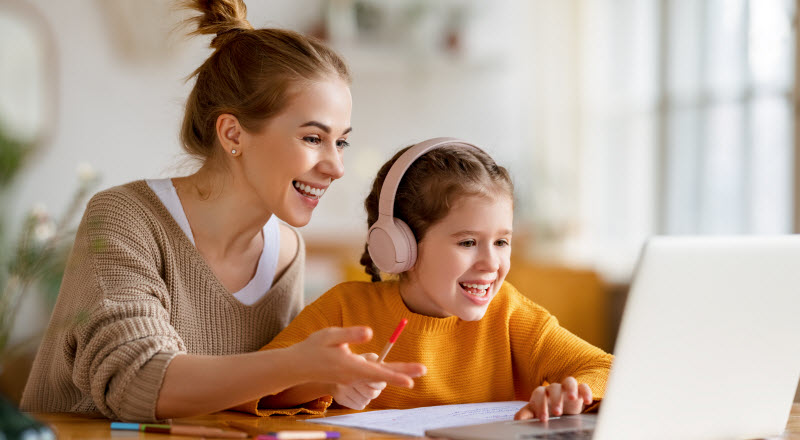 Cairns Paediatric Psychology - Key points helping a child with ADHD Cheerful little schoolgirl in headphones talking to the teacher while studying remotely via laptop at home with happy mom sitting nearby